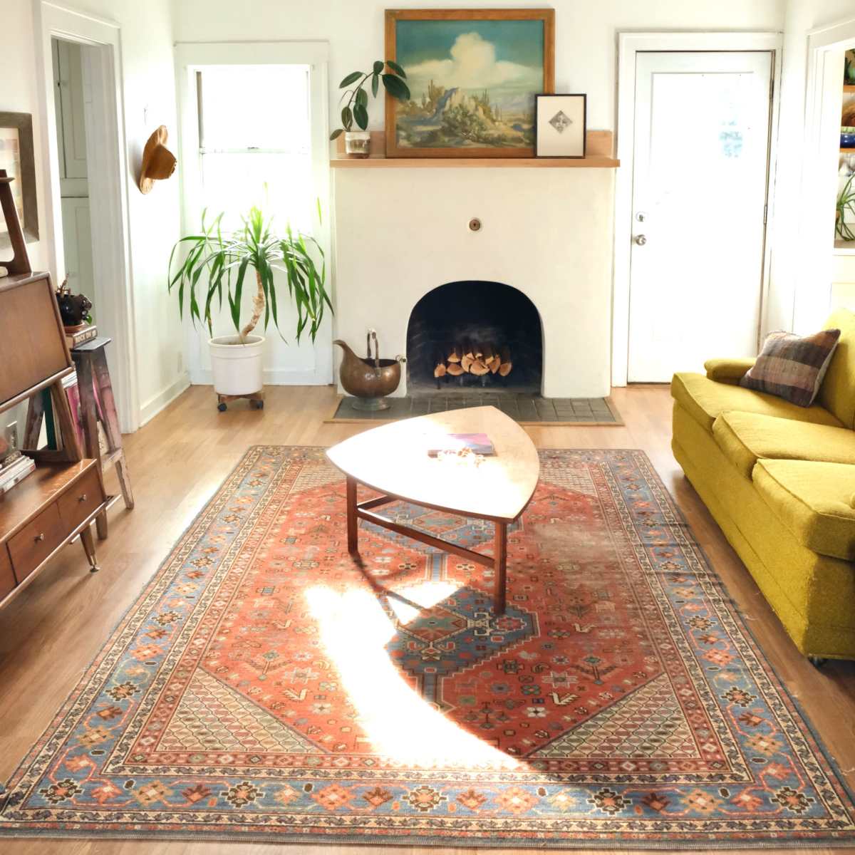 A bright living room features a green sofa, a coffee table on a patterned rug, and a decorative fireplace with a painting above it.
