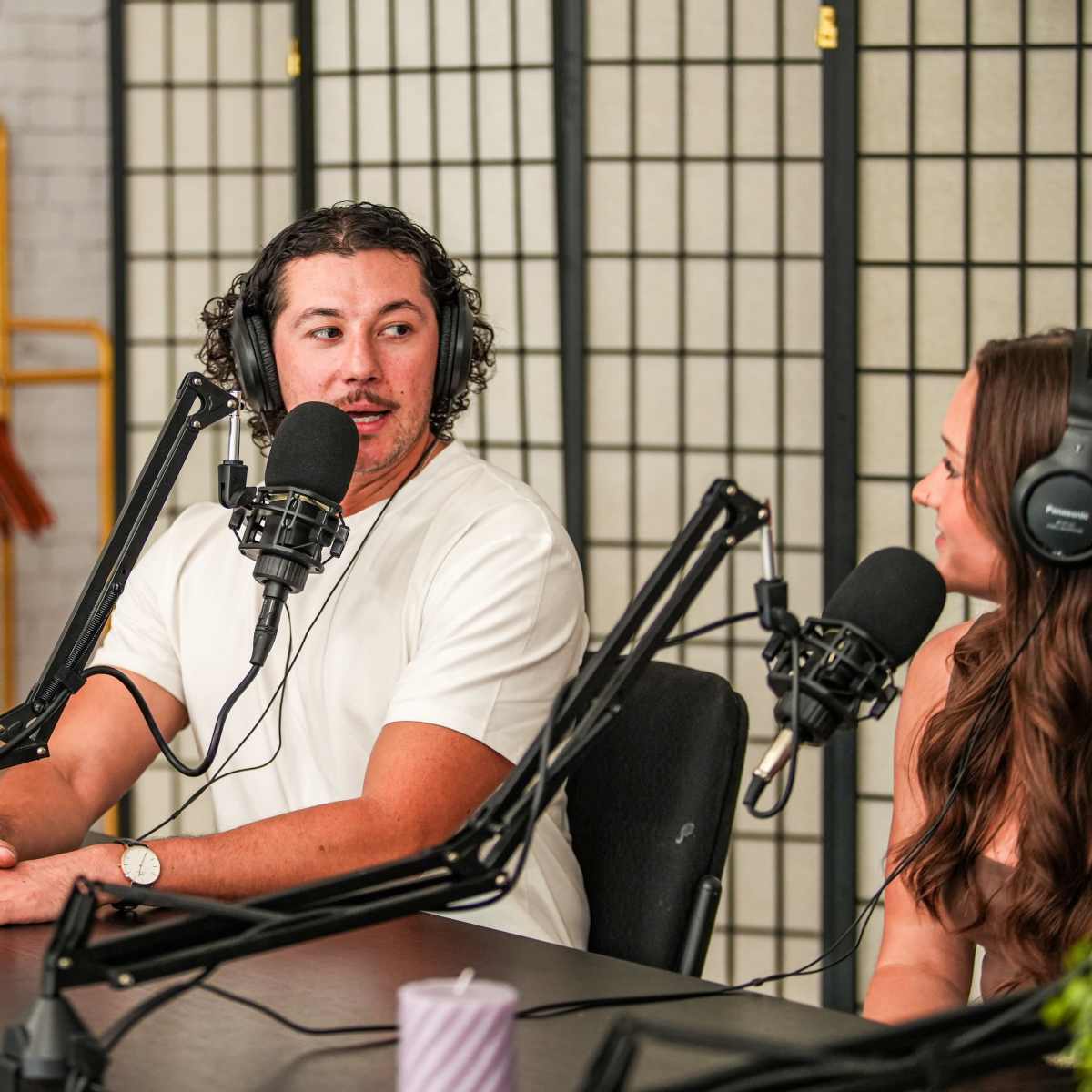 Two people are engaged in a podcast discussion at a table, with microphones in front of them and a potted plant nearby.