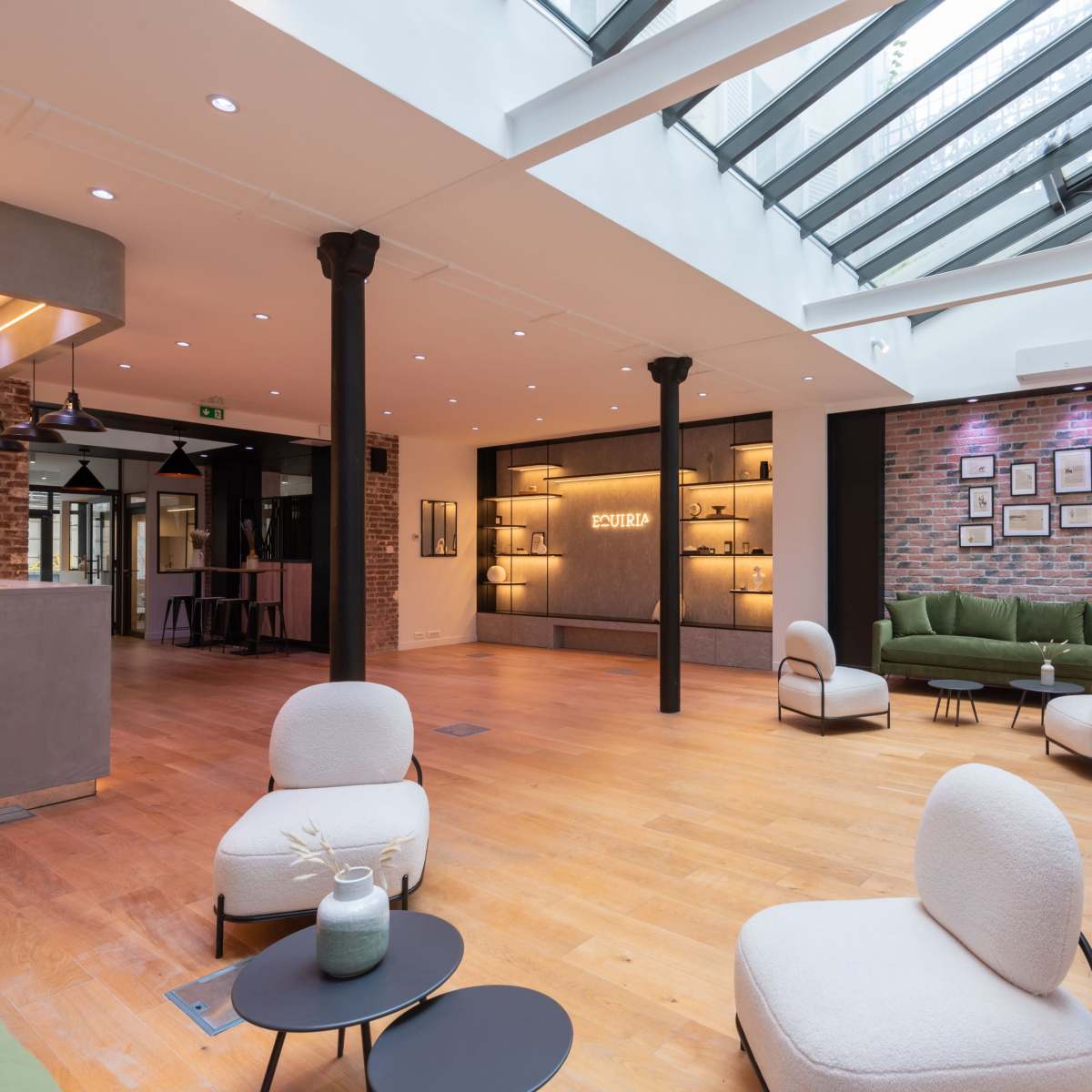 A modern interior space with wooden flooring, featuring green sofas, minimalist chairs, and a glass ceiling that allows natural light to flood in.