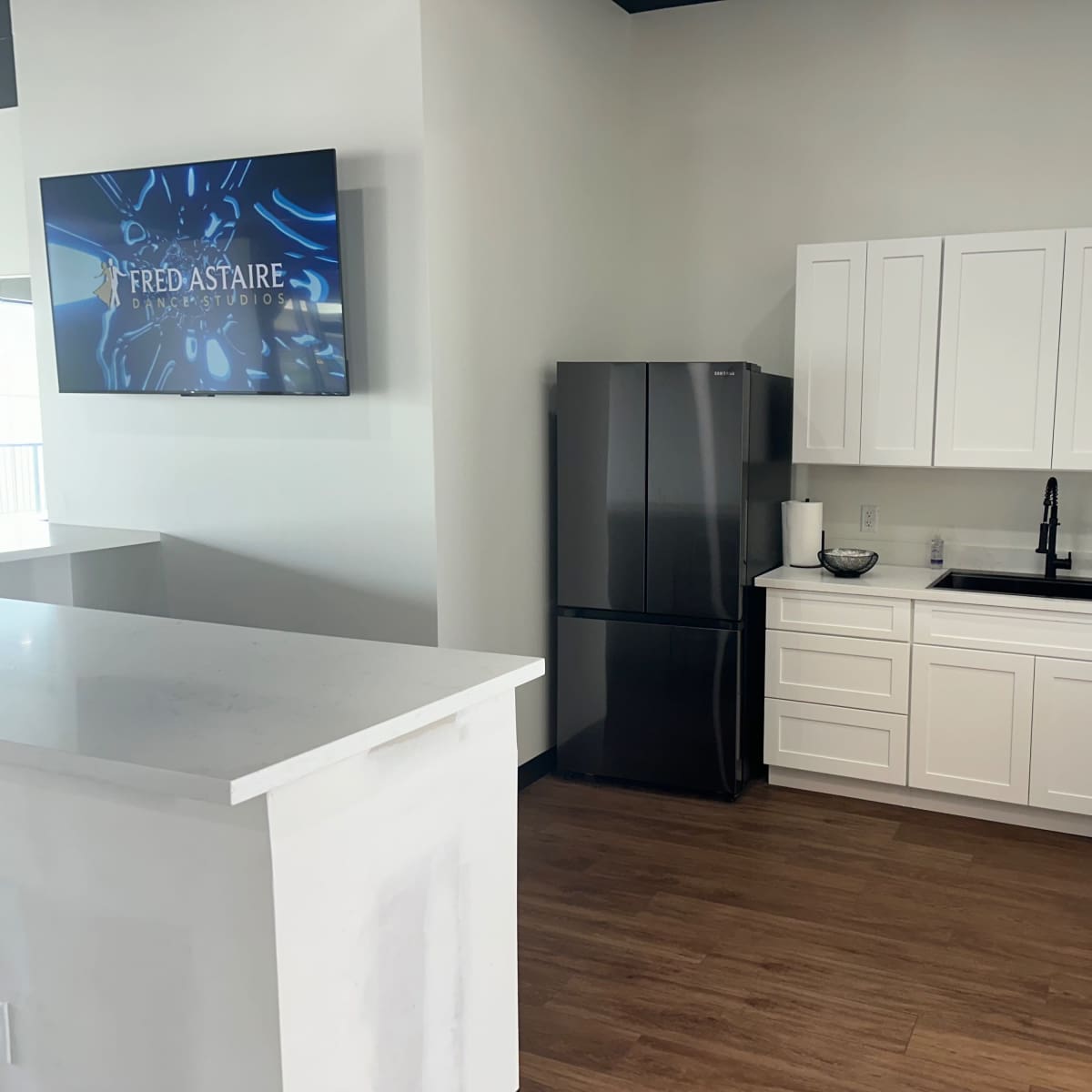 A modern kitchen space with white cabinetry, a dark refrigerator, and a wall-mounted screen displaying the logo of Fred Astaire Dance Studios.