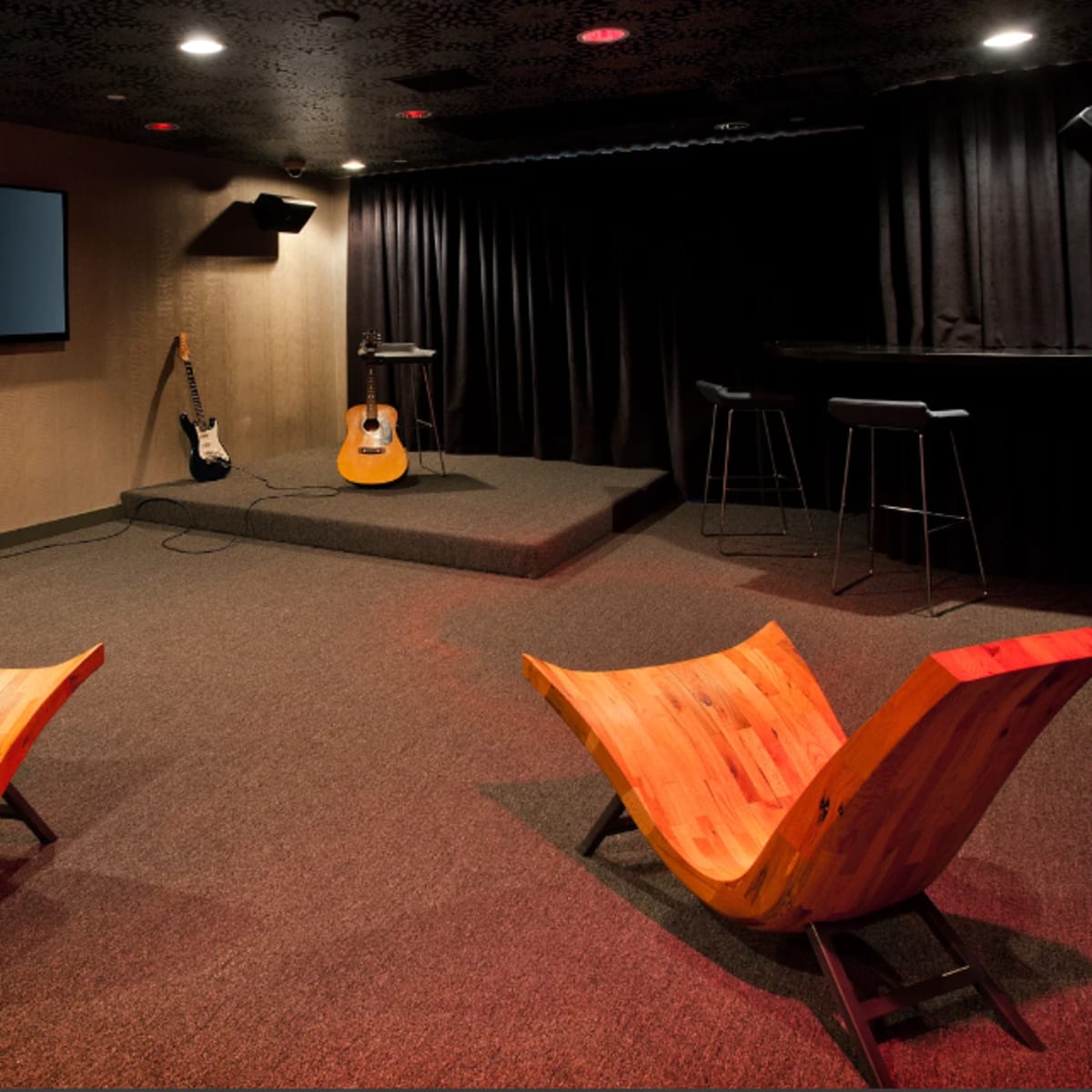 A small performance space with two wooden chairs facing a stage equipped with a guitar, microphone stand, and a TV screen.