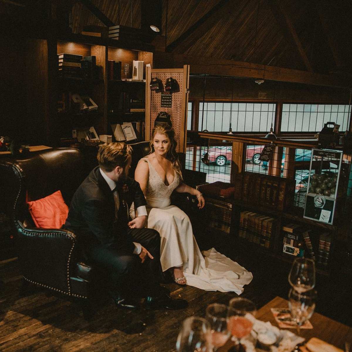 A couple sits together in an intimate setting, surrounded by bookshelves and warm lighting, with glasses on a table nearby.