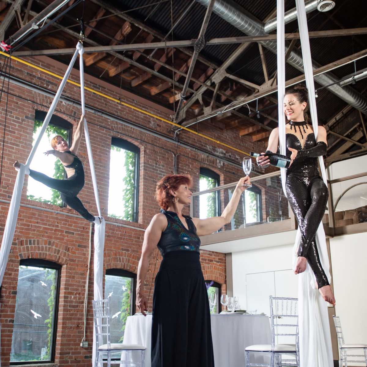 Aerial performers showcase their skills in a spacious, industrial-style venue while a woman in formal attire hands a glass to a performer suspended from silk.