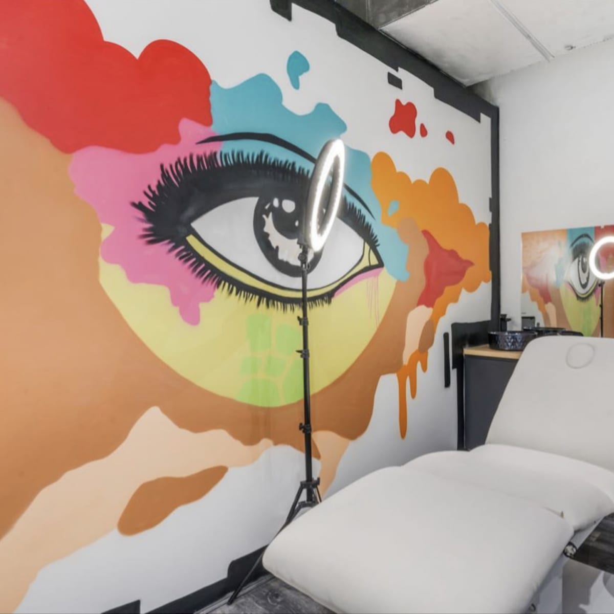 A beauty treatment room featuring a large colorful mural of an eye on the wall, a treatment chair, and a mirror with a ring light.