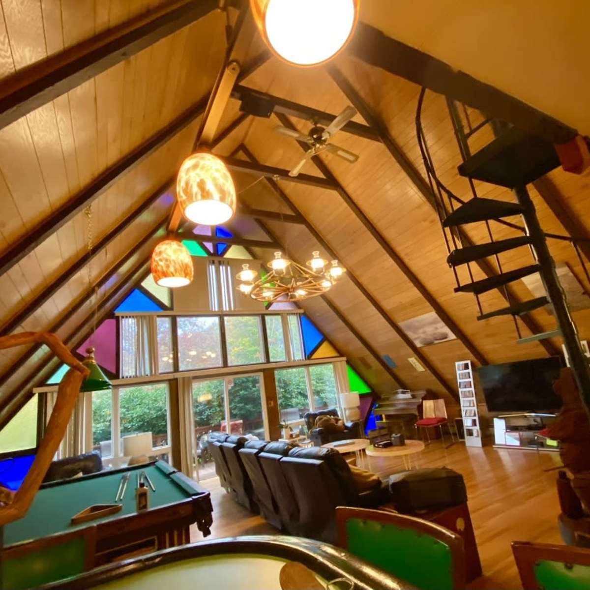 An A-frame living room with wooden beams, a spiral staircase, and colorful stained glass windows overlooking a garden.