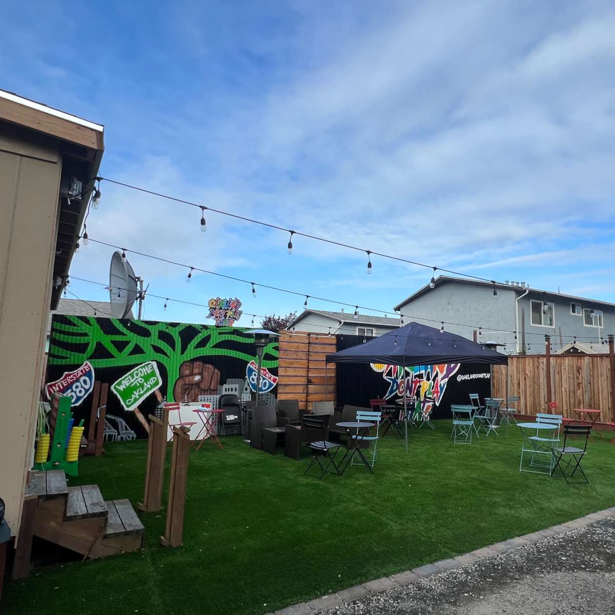 A backyard area with green artificial grass, outdoor furniture arranged around a tent, and decorative mural elements on the walls.