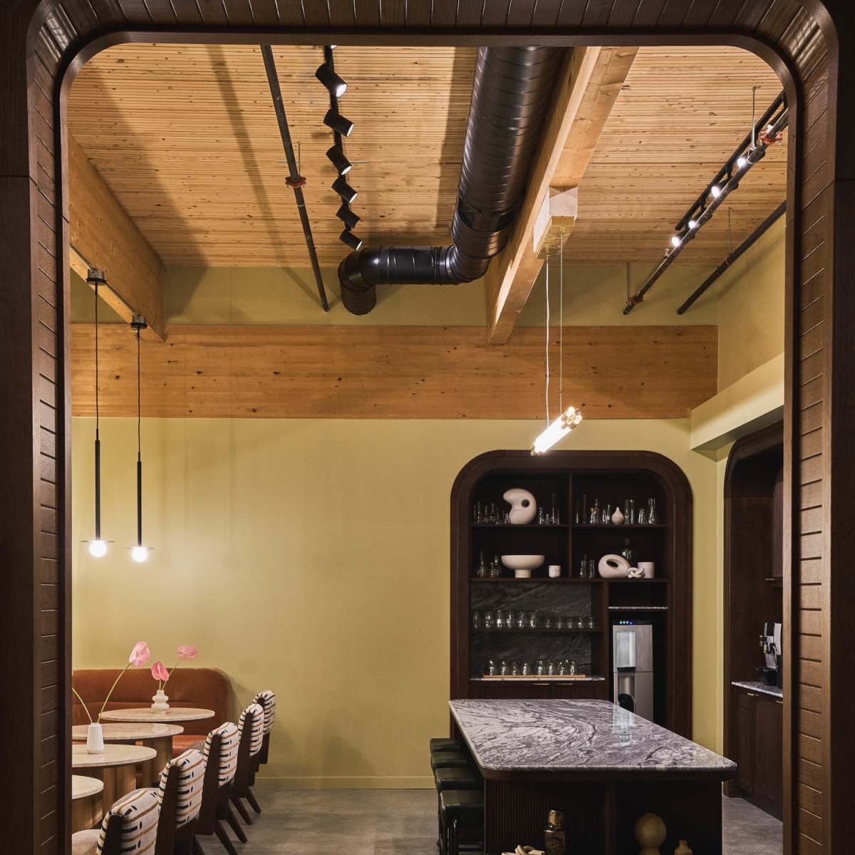 A modern interior featuring a long bar counter with a marble top, dining chairs arranged at tables, and an open kitchen area highlighted by wooden beams and industrial lighting.