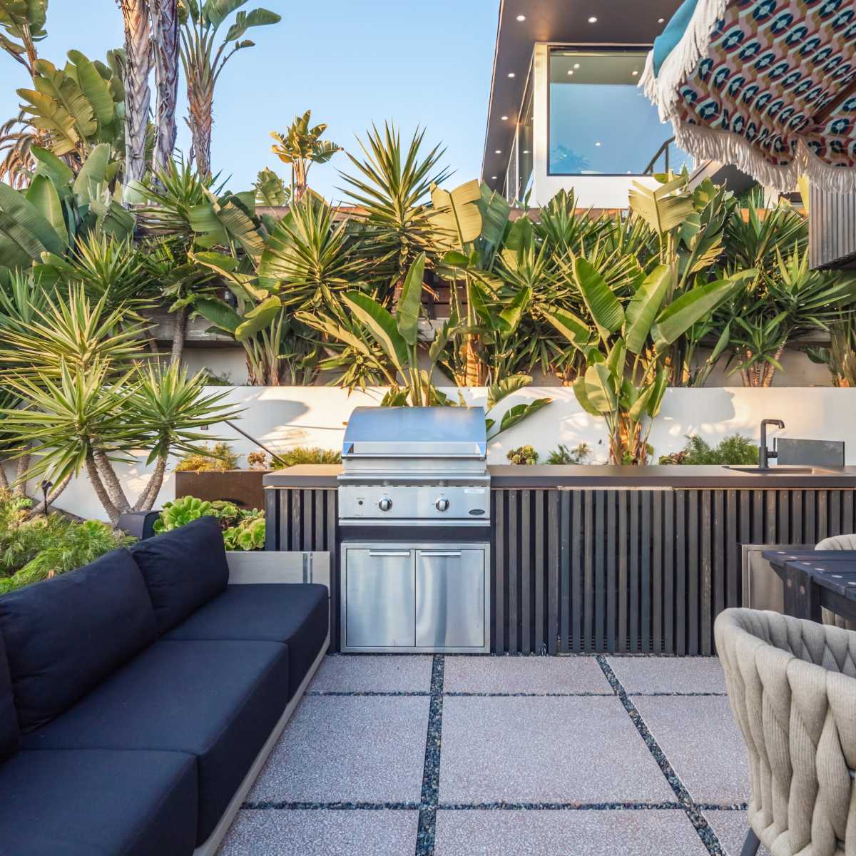 An outdoor kitchen area featuring a stainless steel grill, a dining table, and lush tropical plants surrounding a modern seating arrangement.