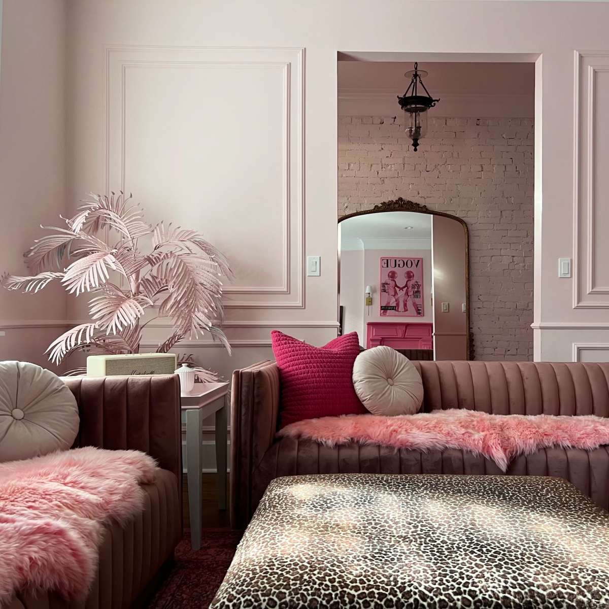 The room features two plush sofas draped with pink fur throws, a leopard-print coffee table, and a large mirror reflecting a stylish decor in soft pastel tones.