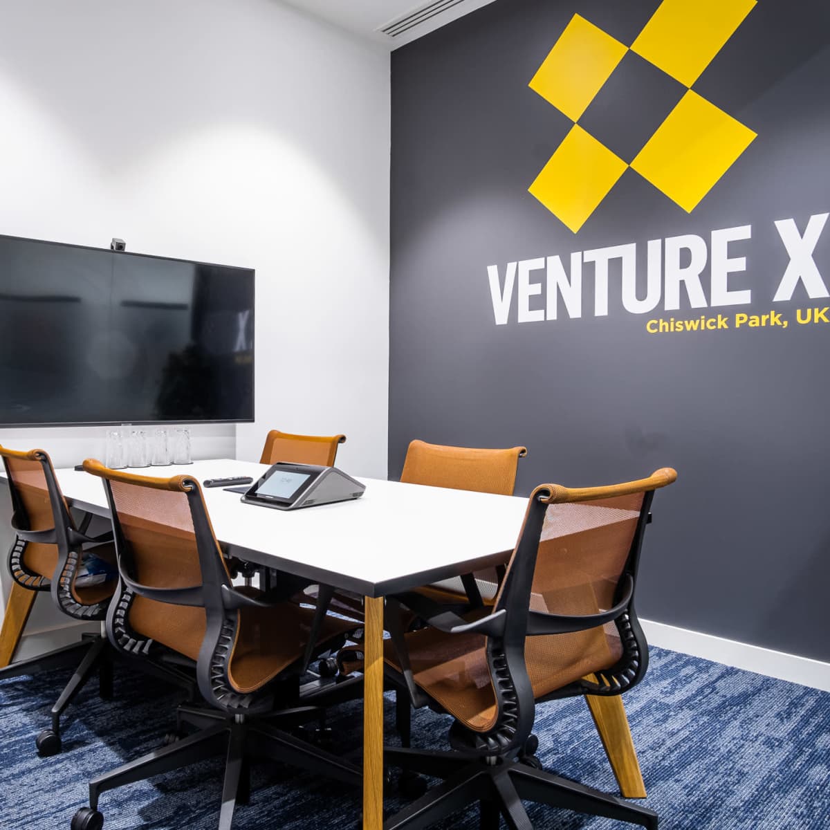 A modern meeting room features a large wall-mounted screen, a white table surrounded by ergonomic chairs, and a backdrop with the Venture X logo.