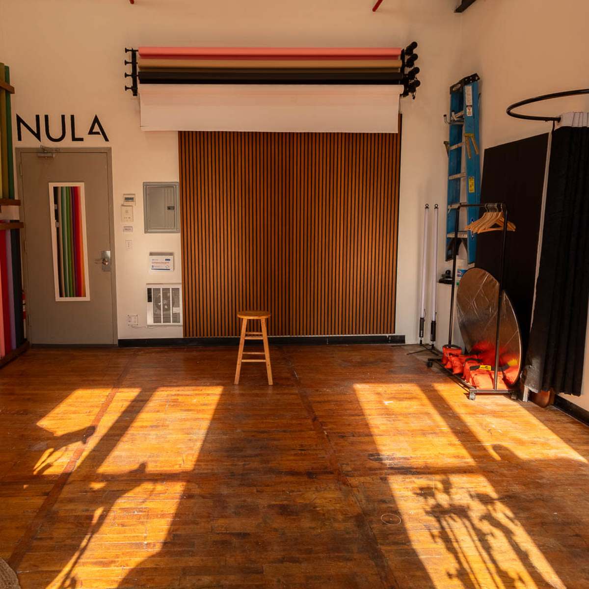 A bright, minimalist studio space featuring colorful vertical stripes on one wall, a wooden feature wall, and a single stool in the center.