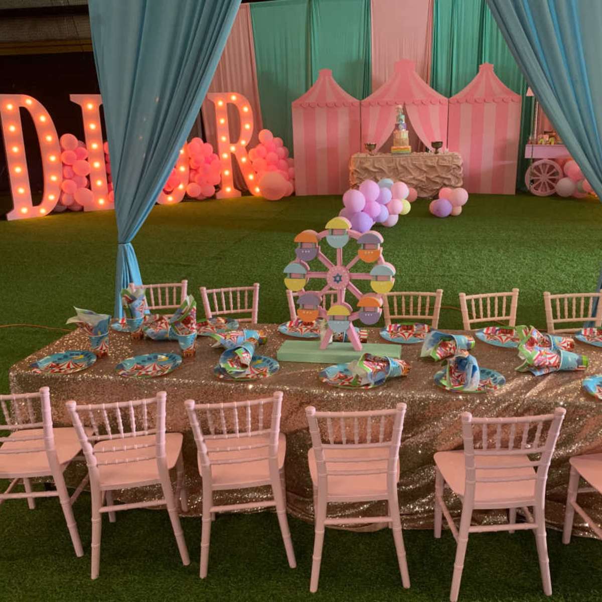A decorated event space features a long table set with colorful plates and a centerpiece, surrounded by pink chairs under draped fabric and balloons.