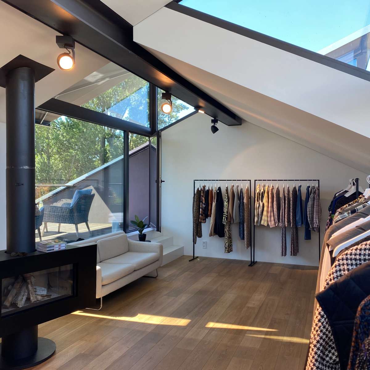 A bright, modern clothing store features a sofa and racks displaying various garments under a sloped ceiling with large windows.