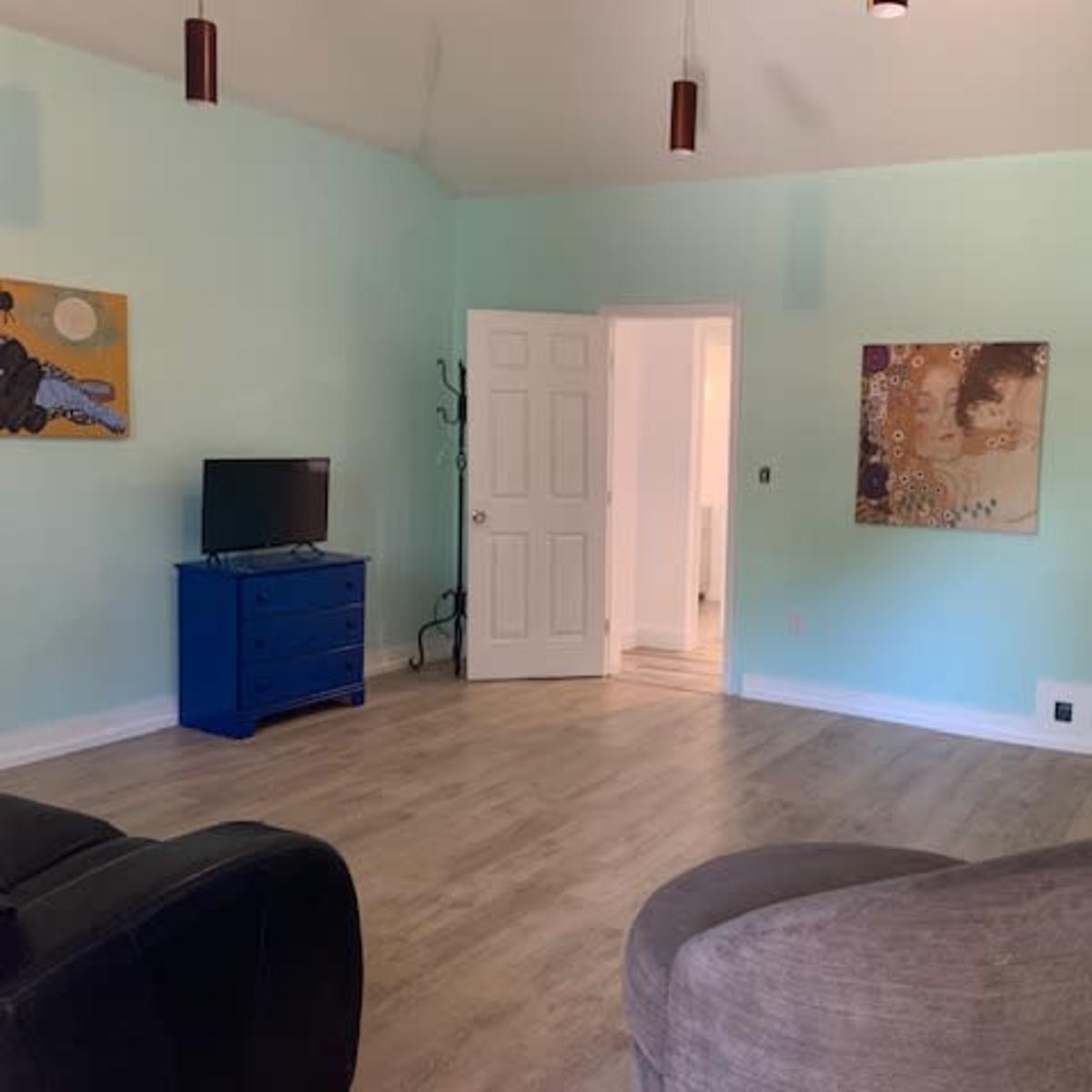 A light blue room with a television on a dresser, a doorway leading to another space, a piece of artwork on the wall, and a modern chair.
