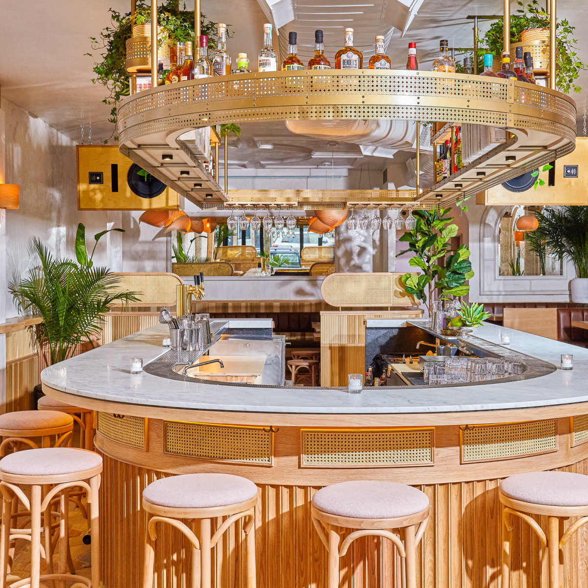 A modern bar area with a circular marble countertop, wooden stools, and decorative plants, featuring shelves stocked with various bottles.