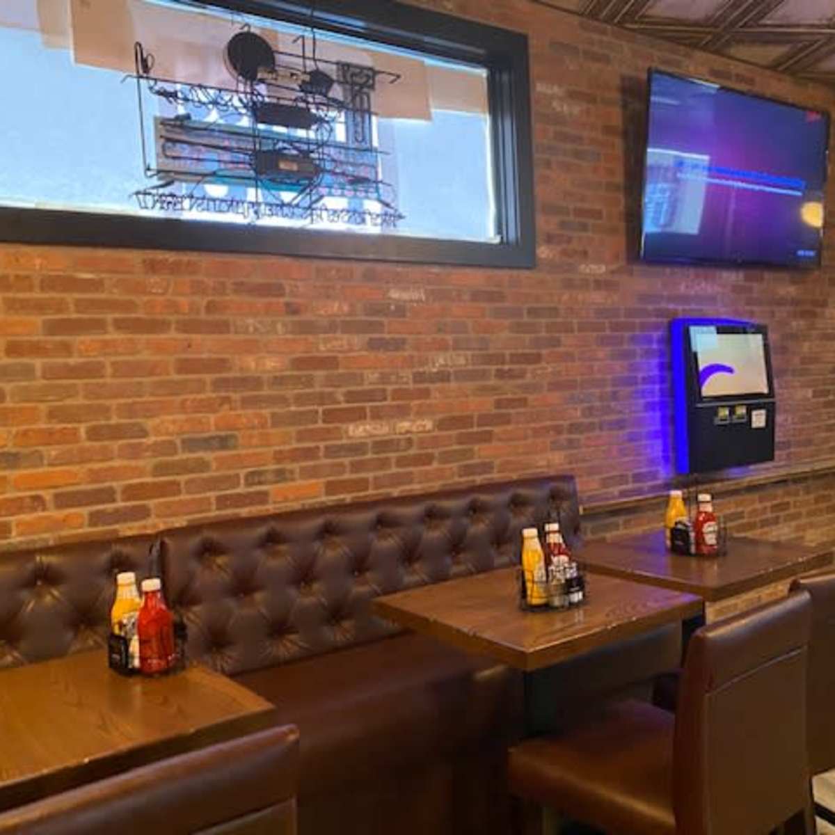 A seating area in a restaurant with brown leather booths, wooden tables, and condiments arranged on the tables against a brick wall.