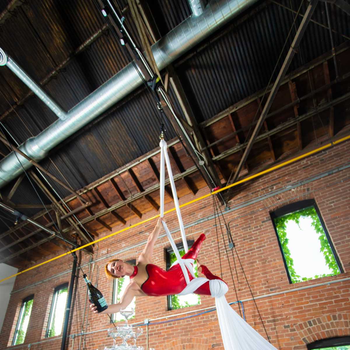 An aerial performer in a red outfit balances gracefully above a glass tower during a festive event in an industrial space.