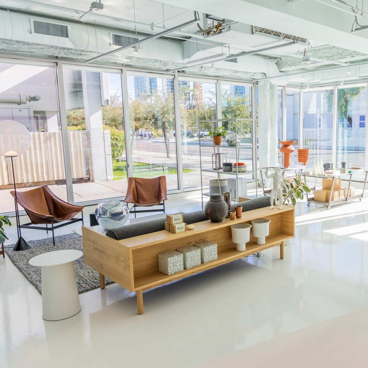 A modern, airy interior space with large windows, featuring various furniture pieces including a wooden bench, lounge chairs, and potted plants.