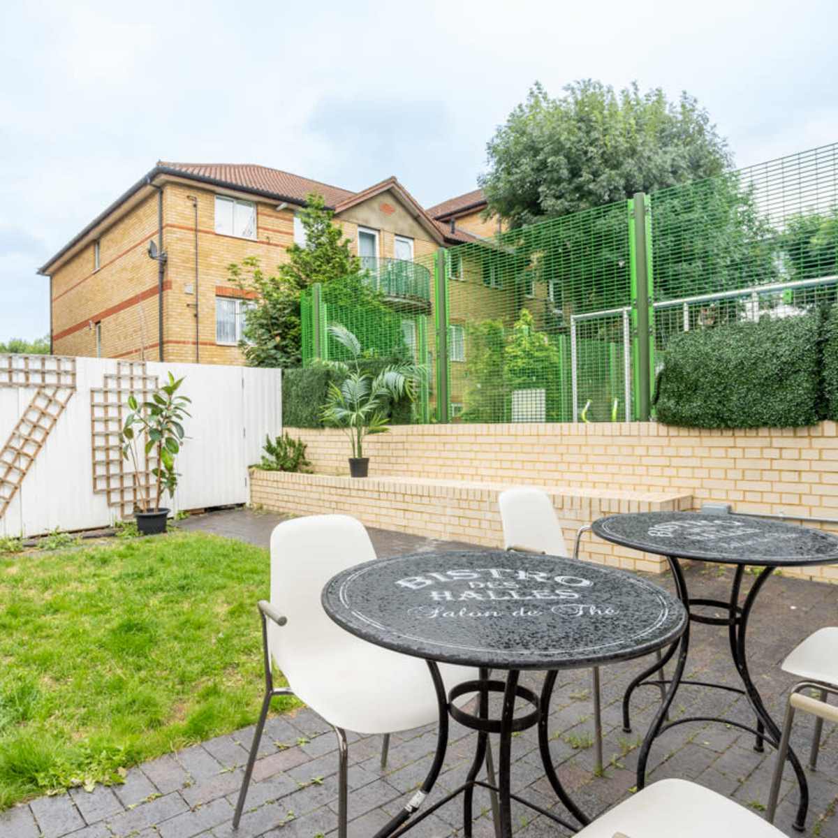 A small outdoor courtyard with two round tables and chairs, bordered by green hedges and residential buildings in the background.
