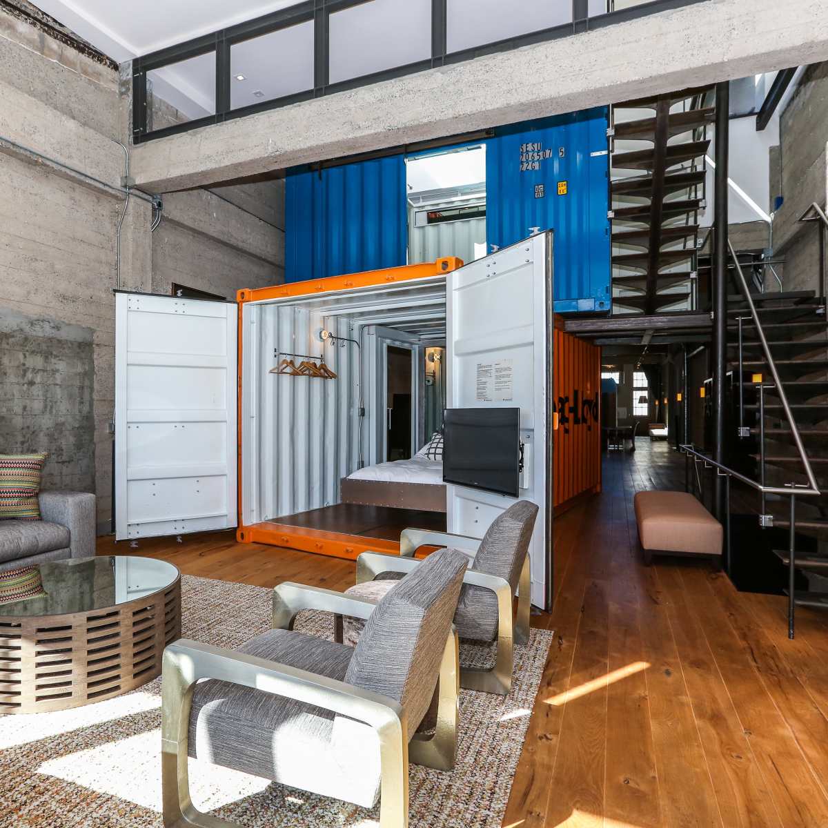 A modern interior space featuring a living area with a seating arrangement, a glass coffee table, and a shipping container used as a bedroom, with a staircase leading to an upper level.
