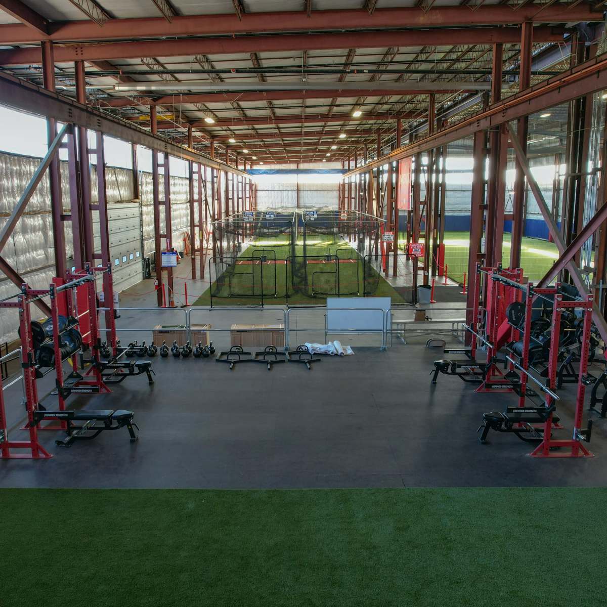 A large indoor sports facility with weightlifting equipment on one side and a training area with turf and batting cages on the other.