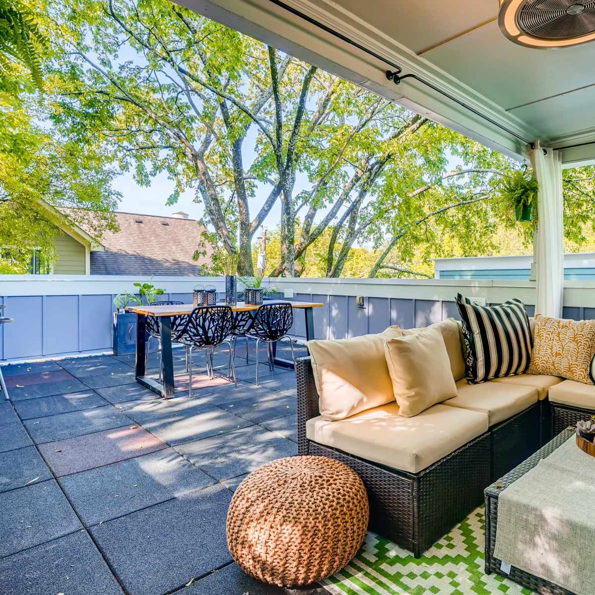 A cozy outdoor patio features a sectional sofa, a coffee table with decorative items, a dining area, and a grill, all surrounded by greenery.