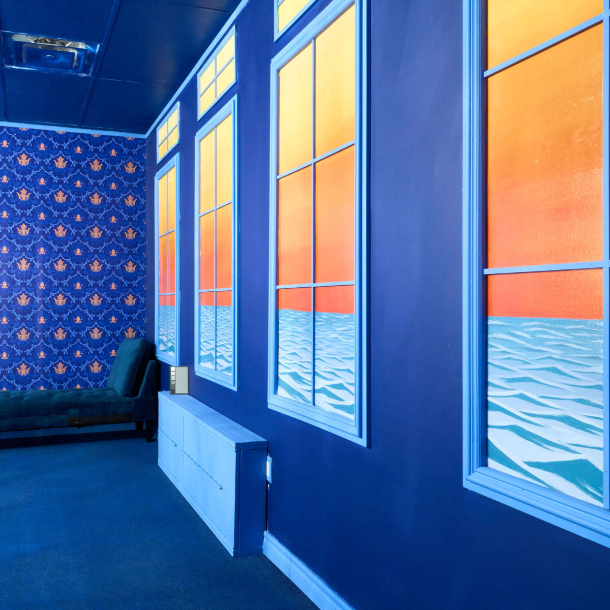 A room with blue walls adorned with a patterned wallpaper, featuring large window murals that depict a sunset over water, alongside a green sofa and a white storage unit.