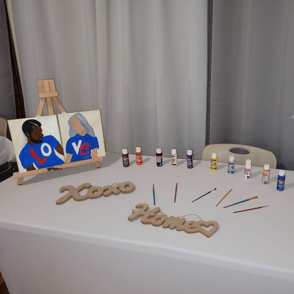 A table displays two painted canvases, multiple bottles of paint, and wooden cutouts spelling "Xoxo," "Home," and "Love."