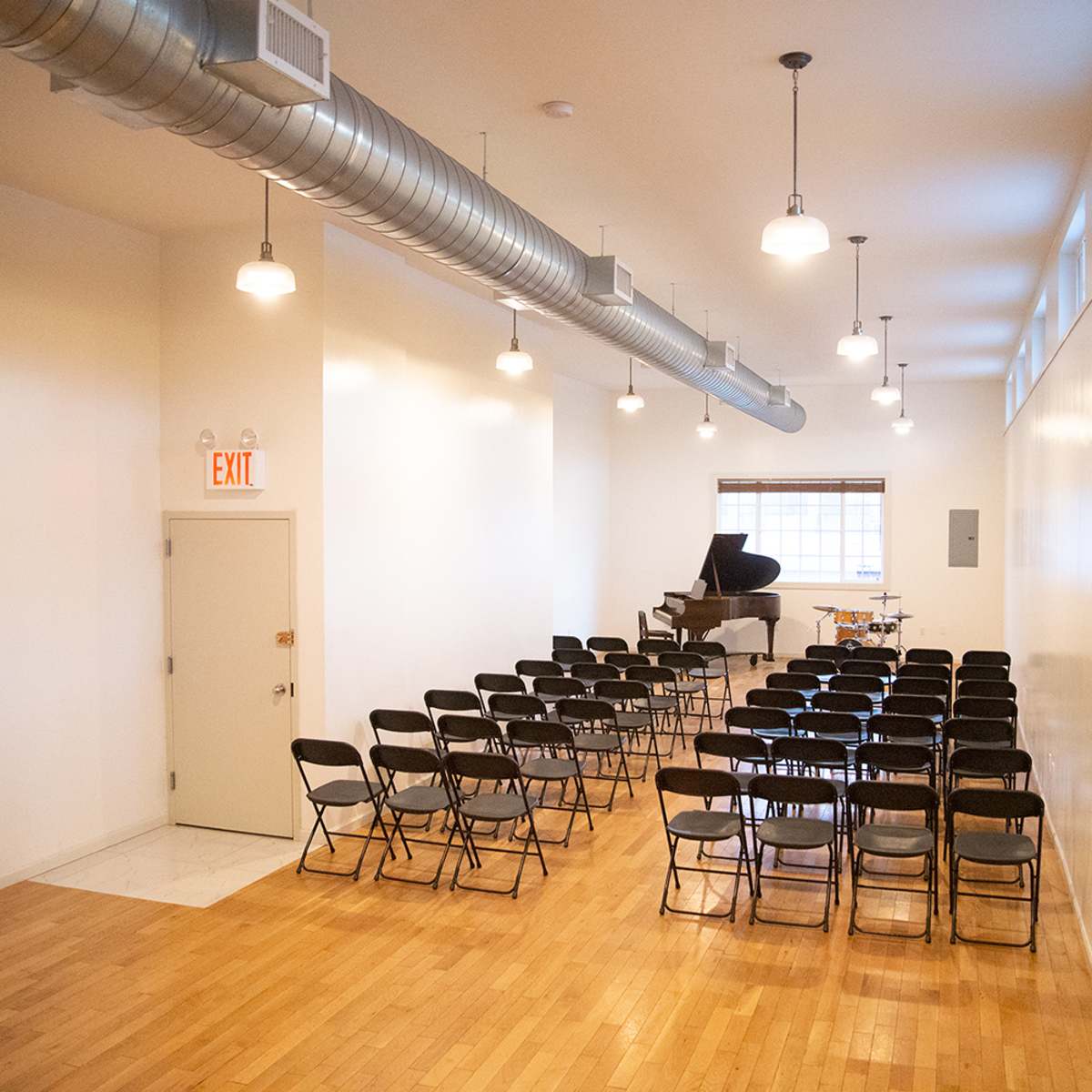 A small performance space features a grand piano and rows of black folding chairs arranged on a wooden floor.