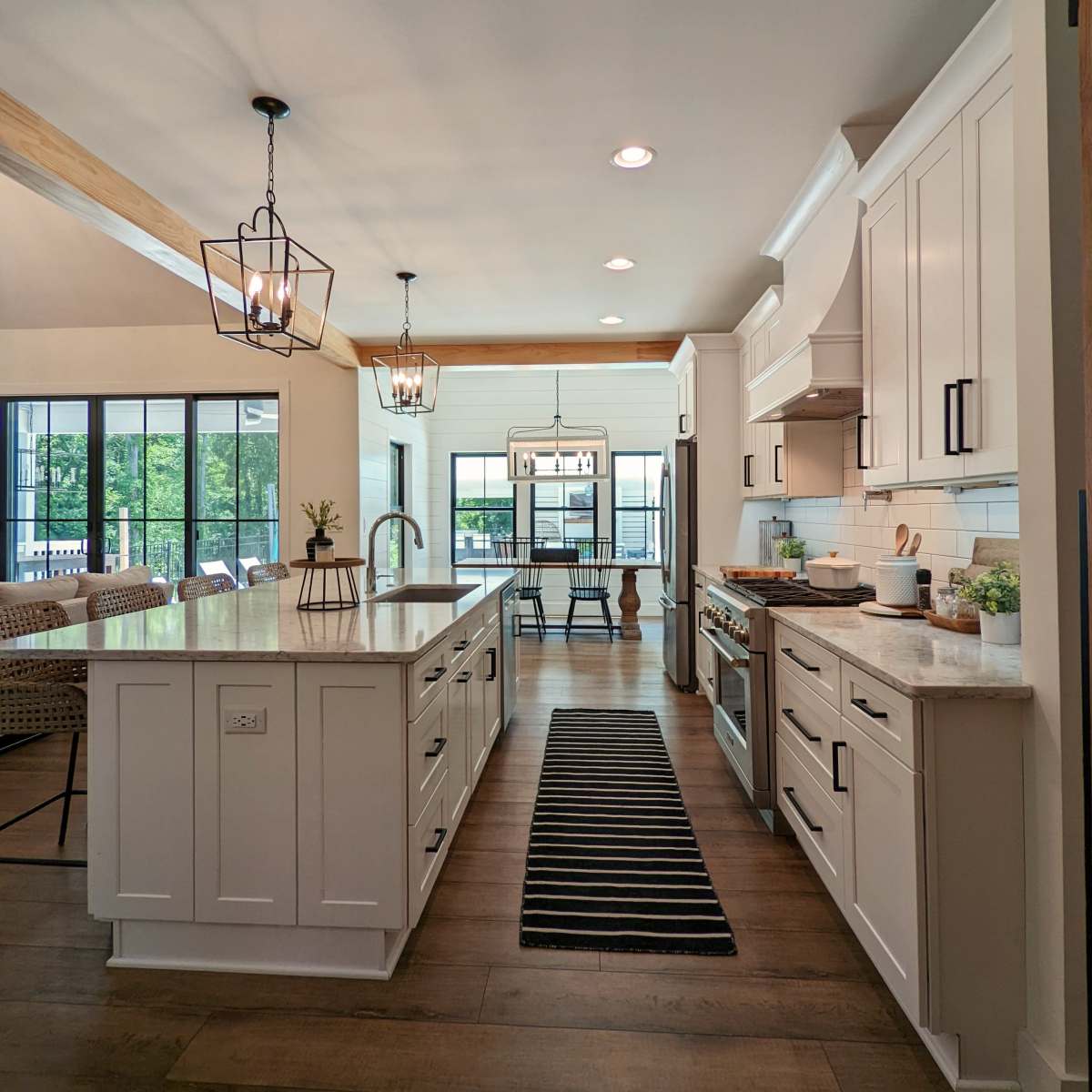 A modern kitchen features white cabinetry, a central island with a sink, and a dining area visible through large windows.