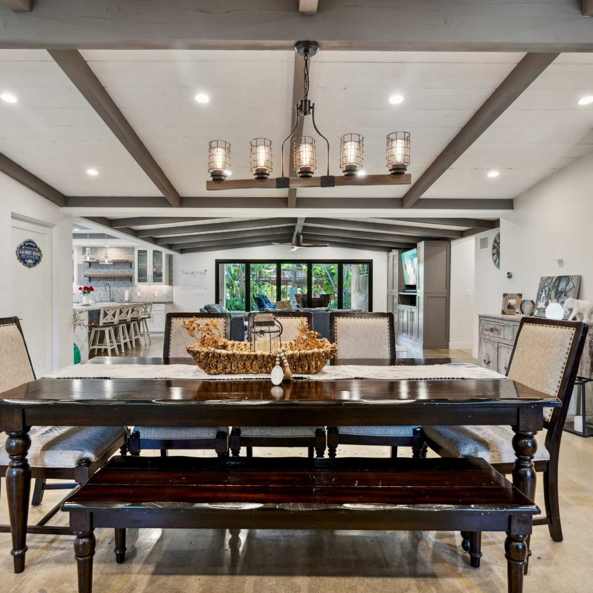 A dining room features a wooden table with a bench and chairs beneath a chandelier, surrounded by white walls and a beamed ceiling.