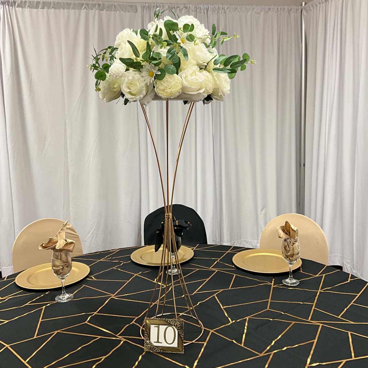 A round dining table is set with a geometric black and gold tablecloth, featuring a tall floral centerpiece and elegantly arranged place settings.