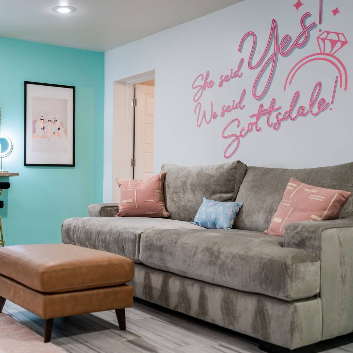 A modern living space features a gray sectional sofa, a brown ottoman, a turquoise accent wall with pink text, and a vanity area with mirrors and bar stools.