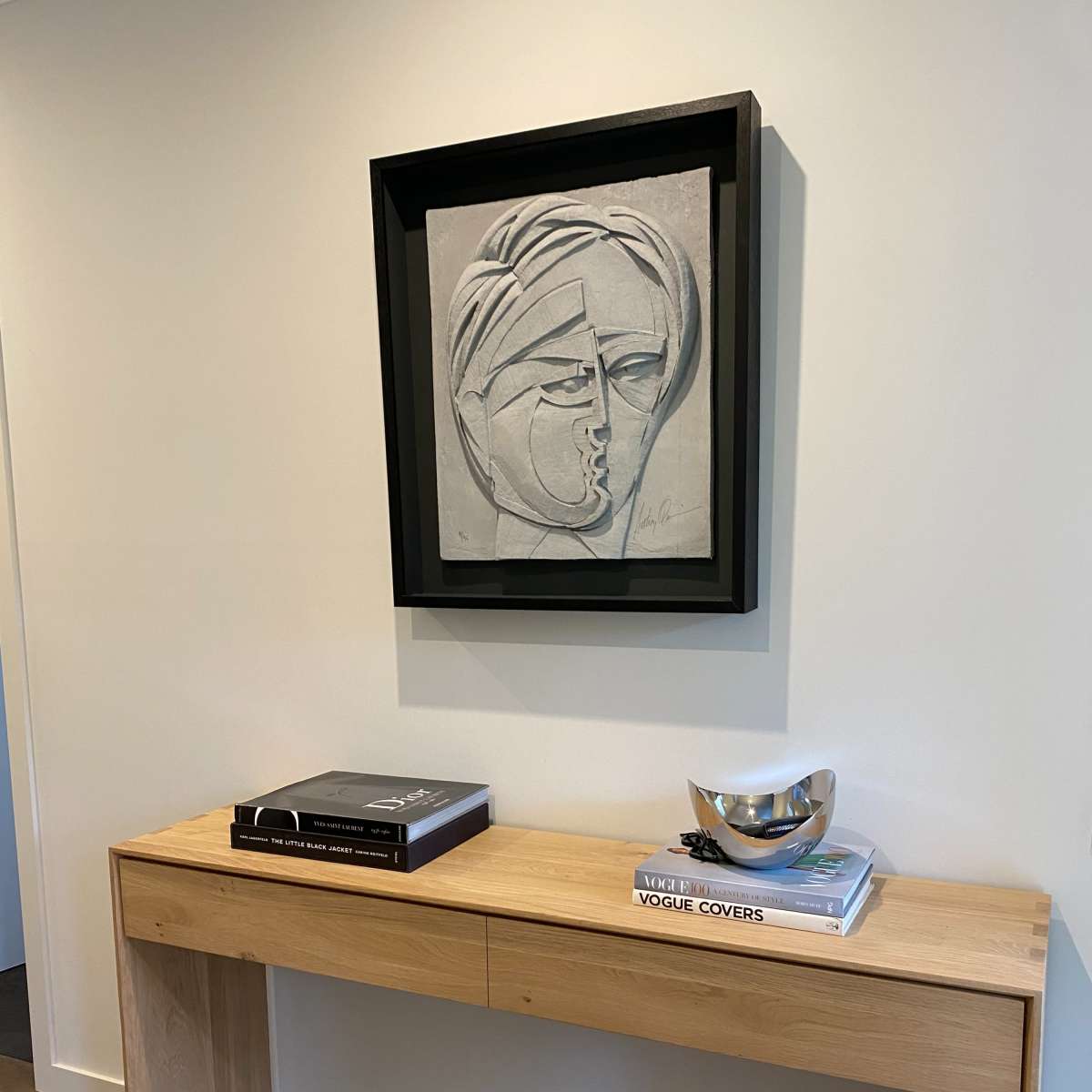 A light-colored wall features a framed abstract artwork above a wooden console table, which holds a stack of books and a decorative bowl.
