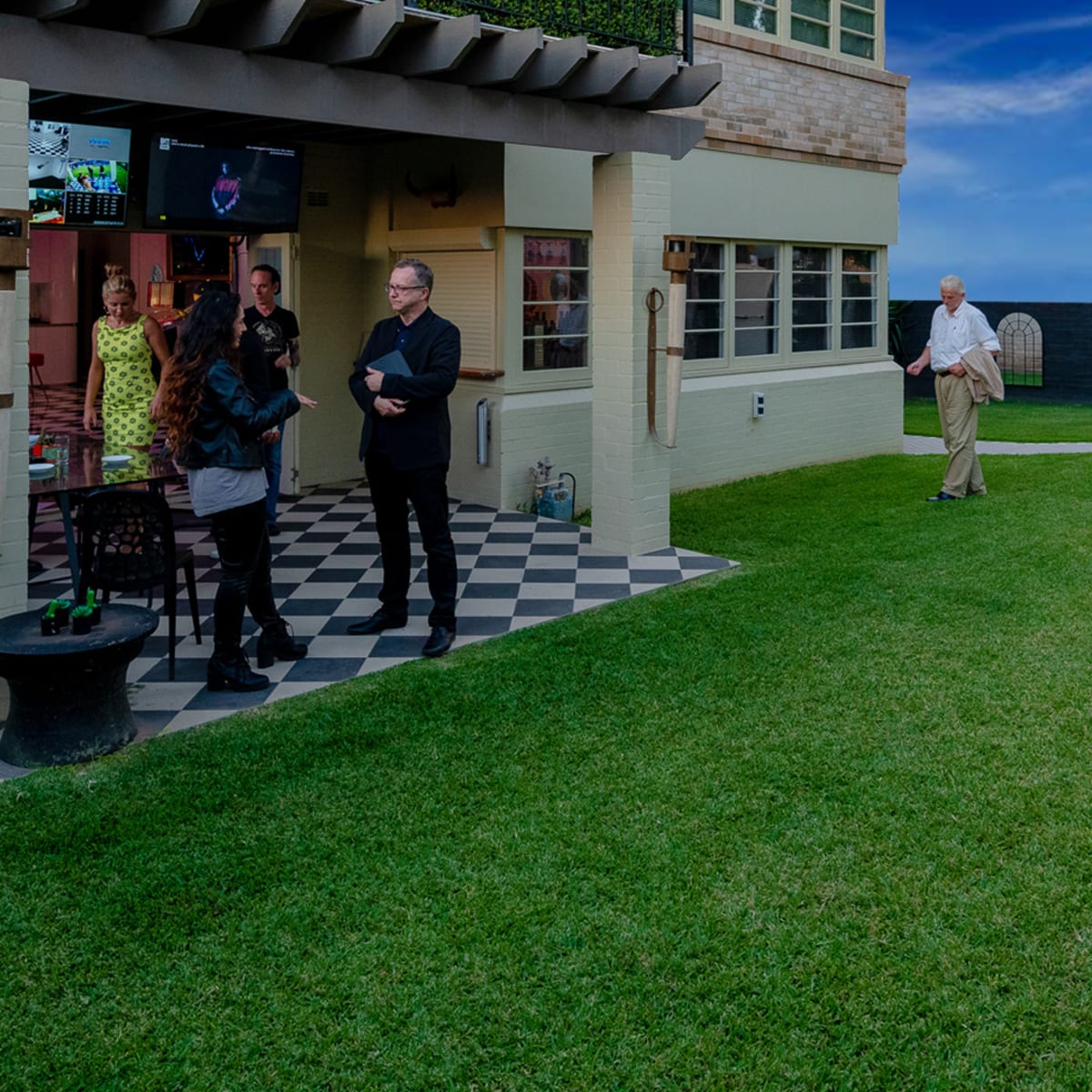 A gathering in a well-manicured lawn with a group of people socializing around a house, a fountain, and a large chair.