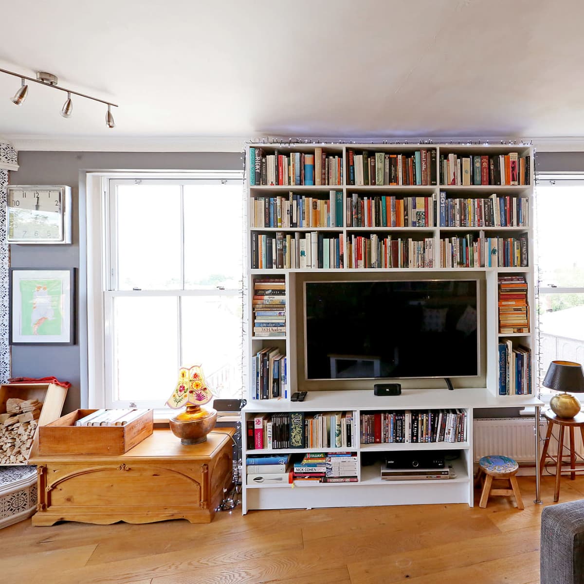 A cozy living room features a large bookshelf filled with books, a flat-screen TV, and windows that provide natural light.