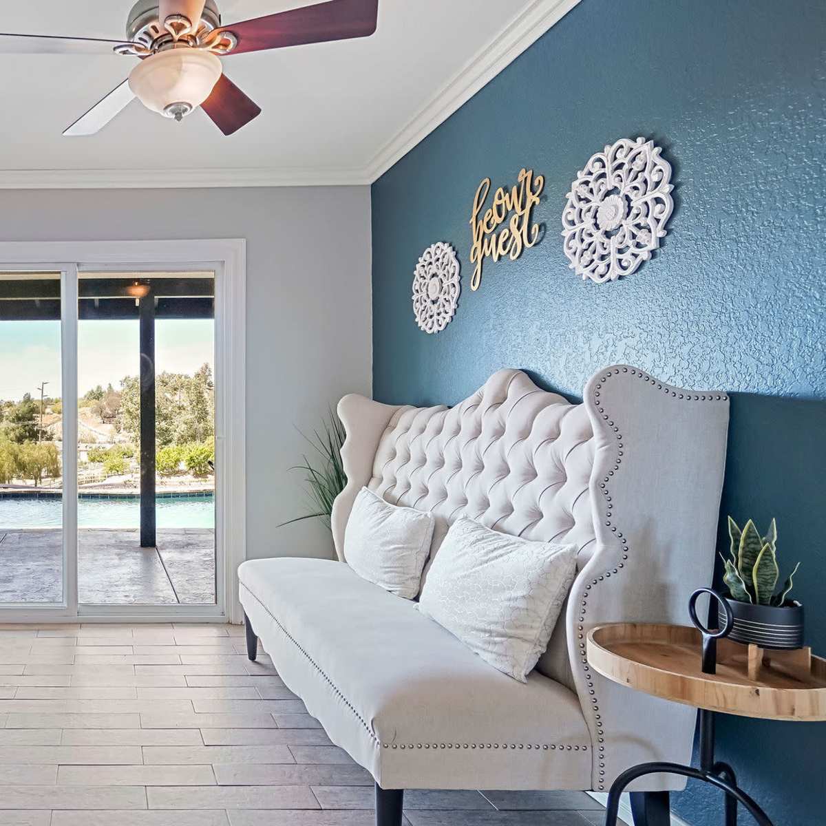 A light-colored living room with a tufted bench against a blue accent wall, leading to large glass doors that open to a view of an outdoor pool area.