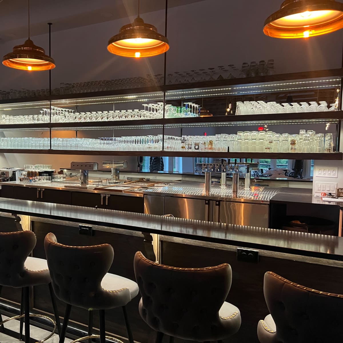 A modern bar area with a sleek counter and rows of glassware displayed on illuminated shelves.