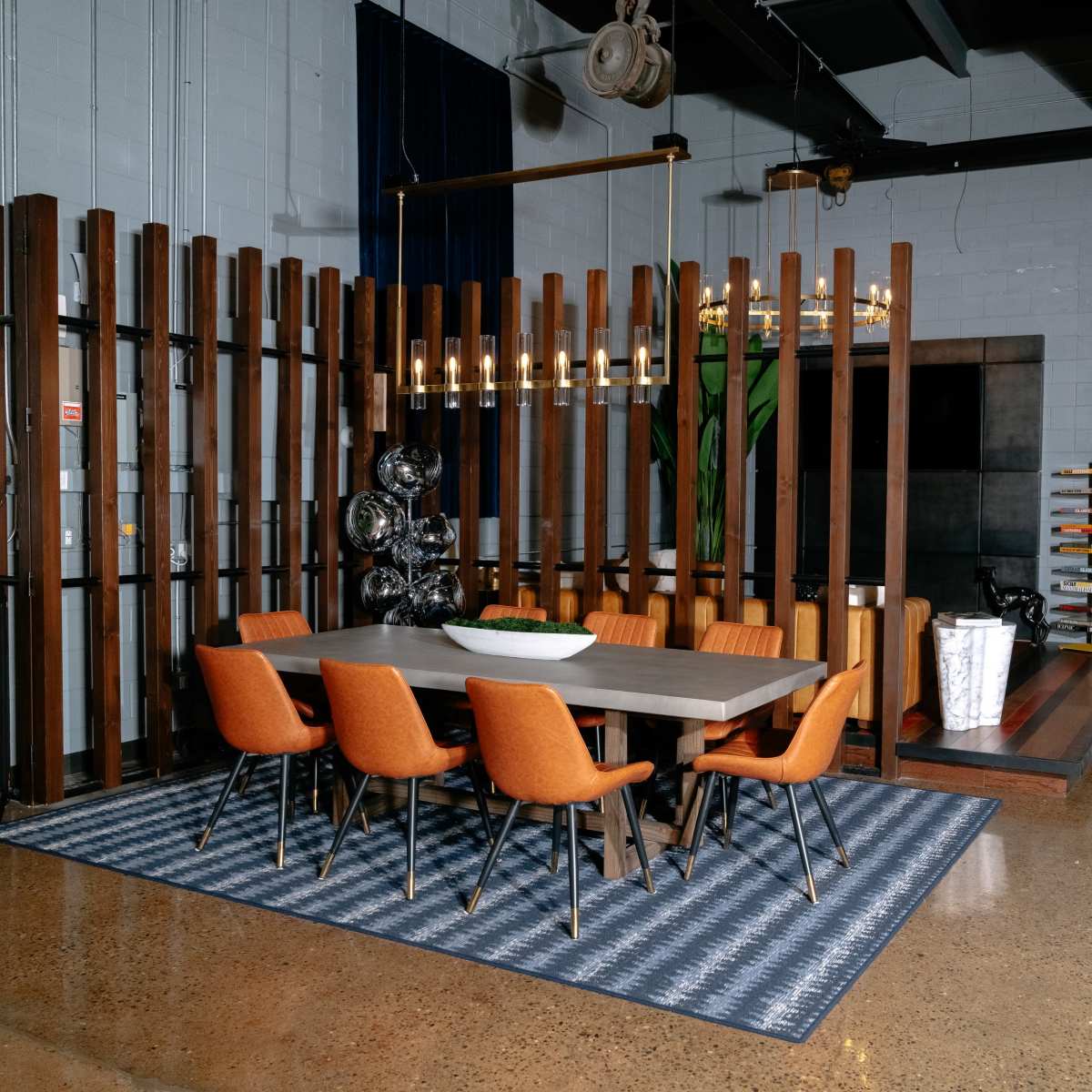 A modern dining area with an eight-seat table surrounded by orange chairs, a patterned rug, and a backdrop of wooden slats.