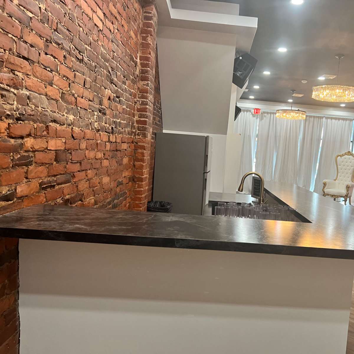 A modern bar area featuring a black countertop, exposed brick walls, and elegant chandeliers overhead.