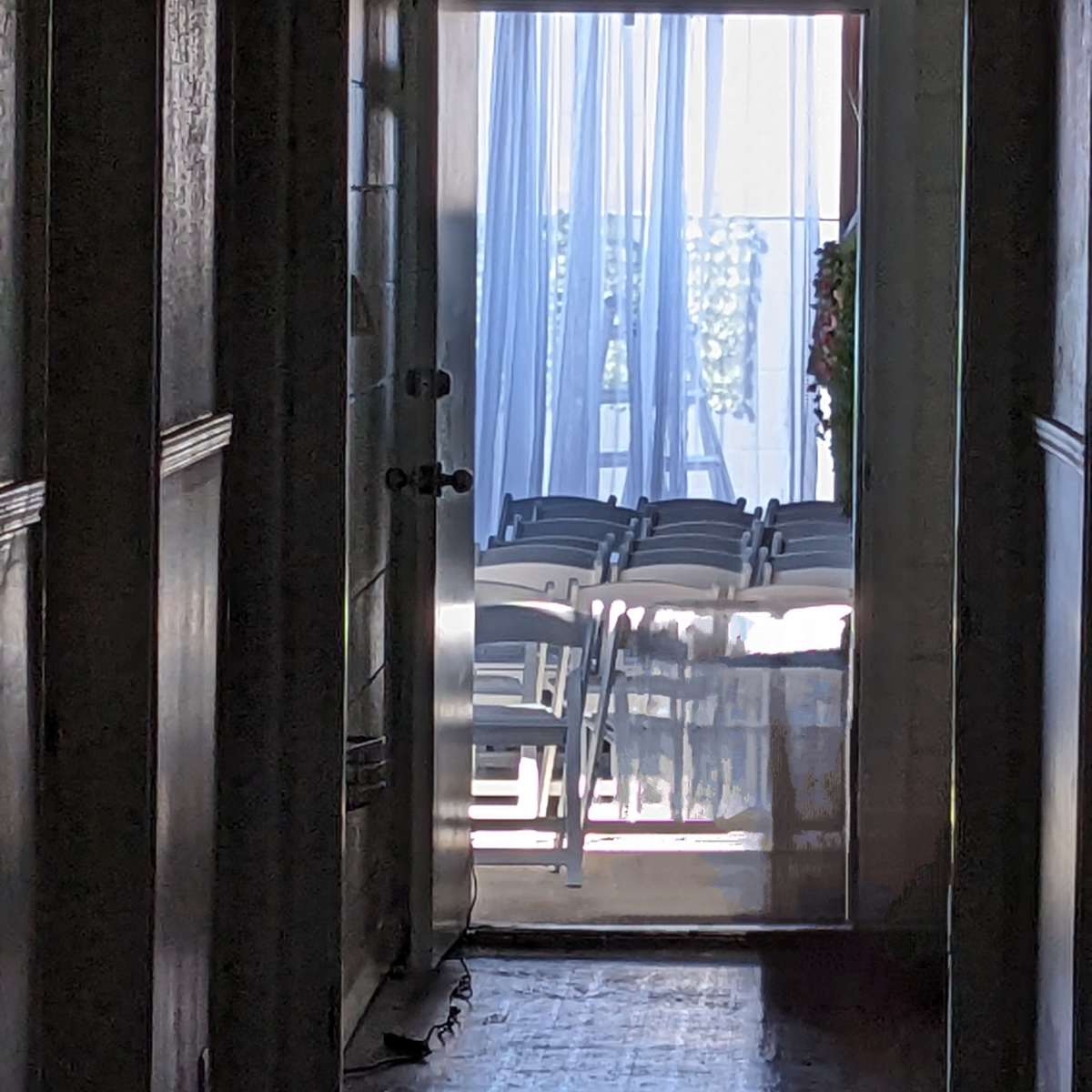A hallway leads to an open door, revealing a room with rows of chairs and sheer curtains in the background.