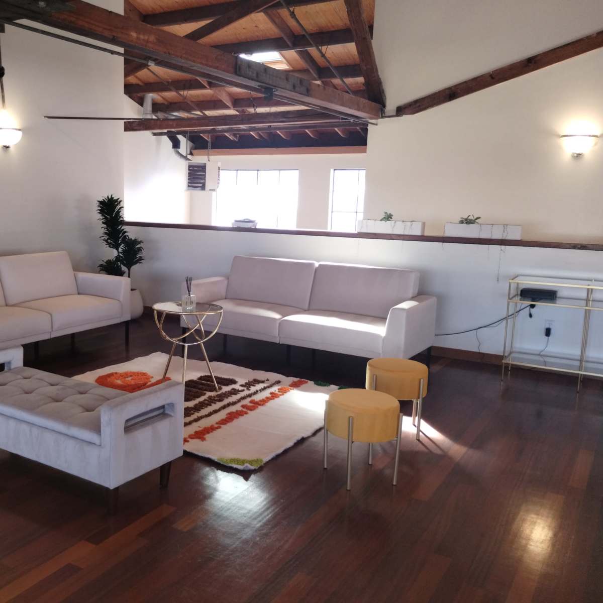 A spacious living area with two sofas, a coffee table, a patterned rug, and a few small stools, all set against a backdrop of wooden beams and large windows.