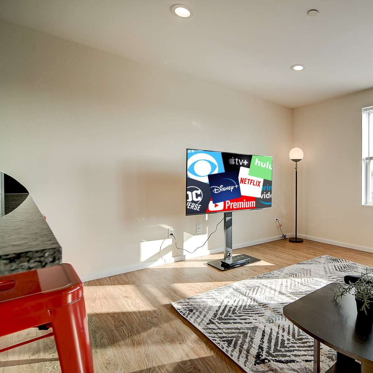 A living room features a television displaying various streaming service logos, with a couch and a lamp situated beside large windows.