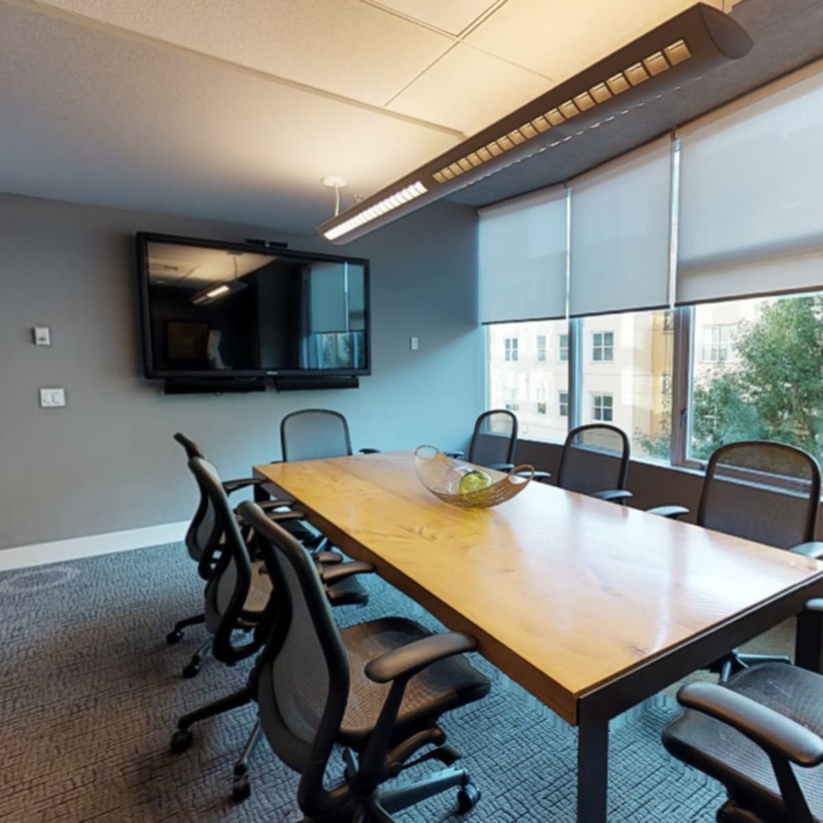 A conference room featuring a long wooden table surrounded by black office chairs and large windows illuminating the space.