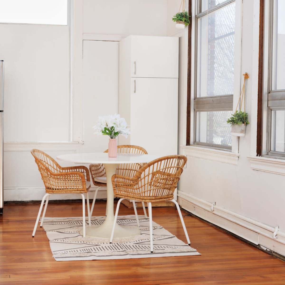 A kitchen area features a round table with two woven chairs, a stainless steel refrigerator, and large windows allowing natural light to enter.