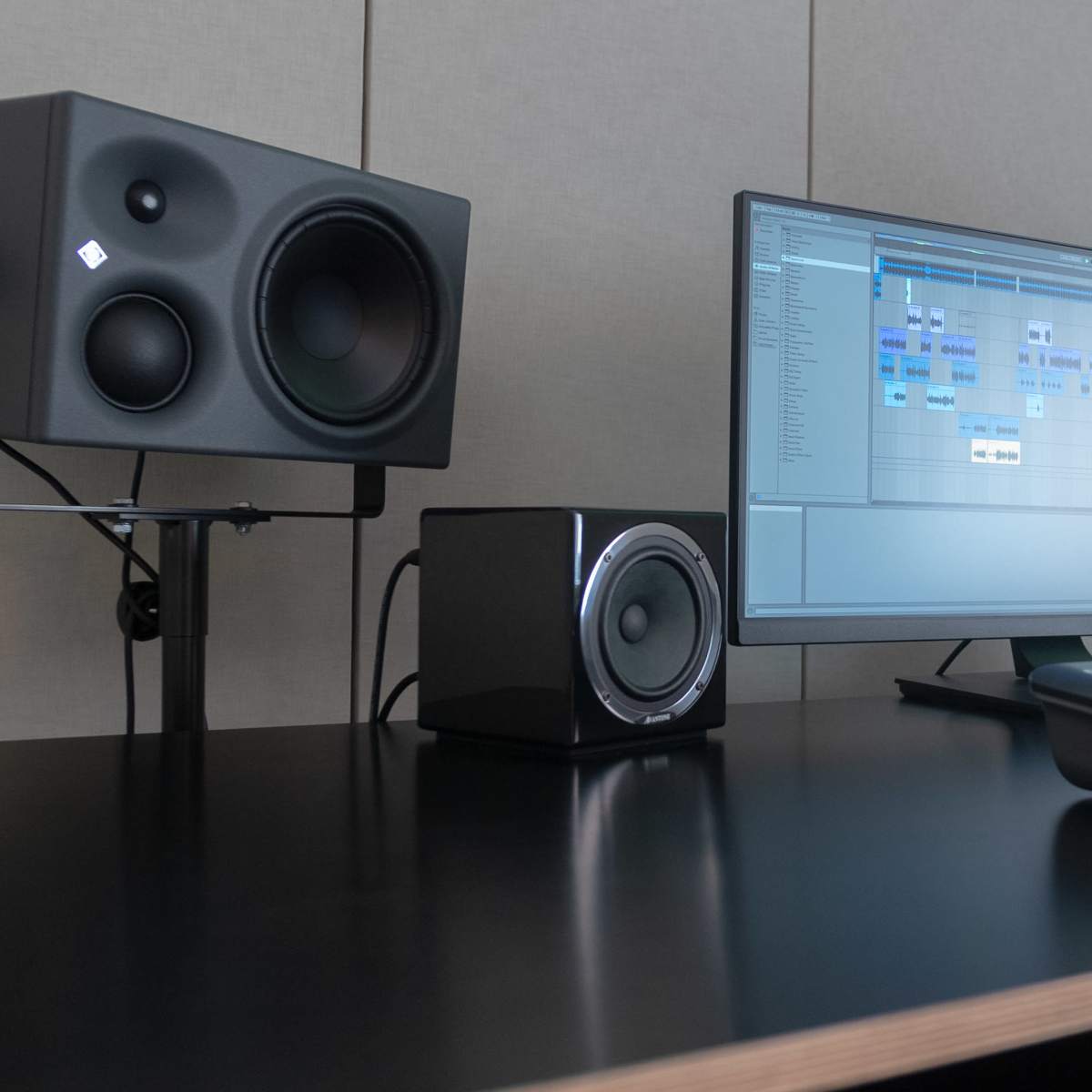 A computer monitor displays audio editing software, flanked by two studio monitors on stands and a keyboard controller on a black desk.