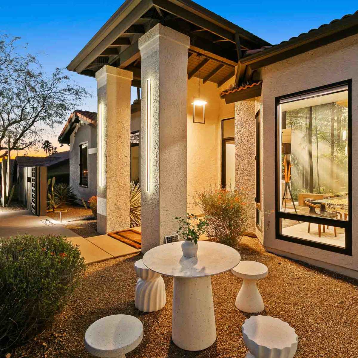 A modern outdoor seating area features a round table and sculptural stools amidst a desert landscape with a backdrop of a contemporary home at sunset.
