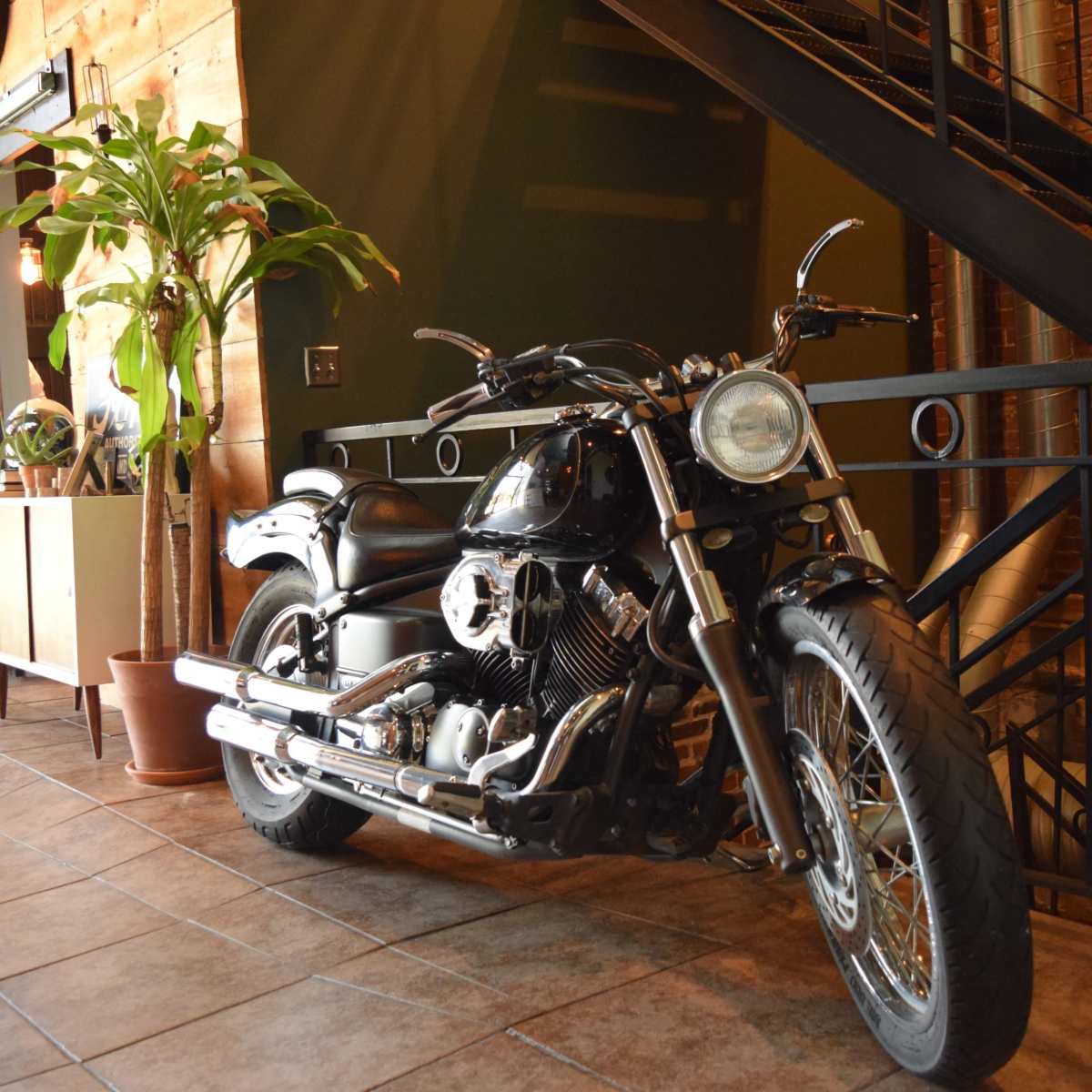 A motorcycle is parked beside a potted plant in an industrial-style interior with brick walls and tile flooring.