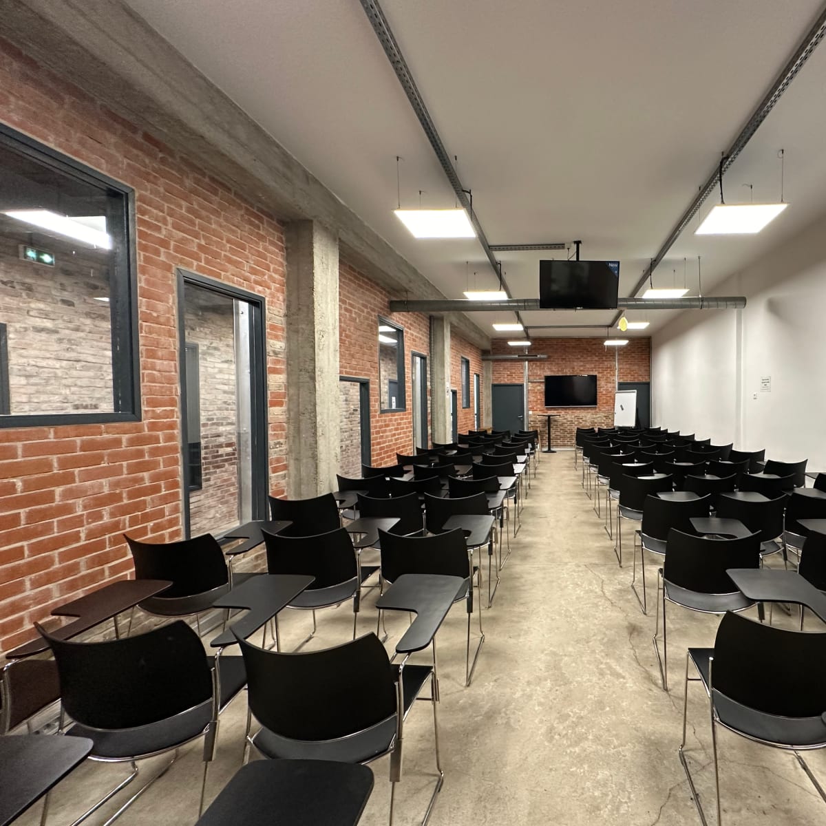 A classroom setup with rows of black chairs arranged facing a screen in a room with exposed brick walls and concrete flooring.