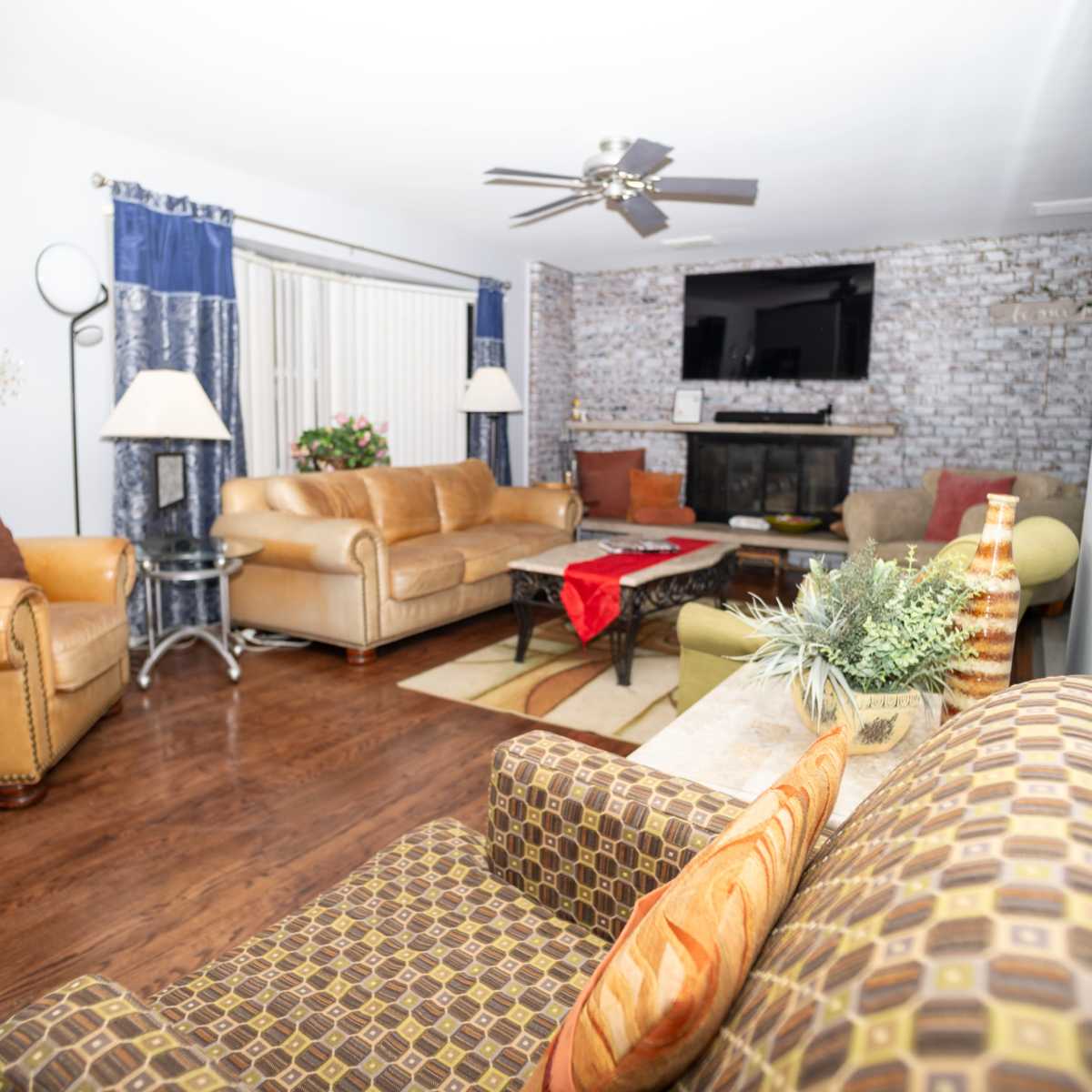 The living room features a combination of brown leather sofas, a patterned armchair, and a decorative coffee table, with a stone fireplace and a flat-screen TV on the wall.